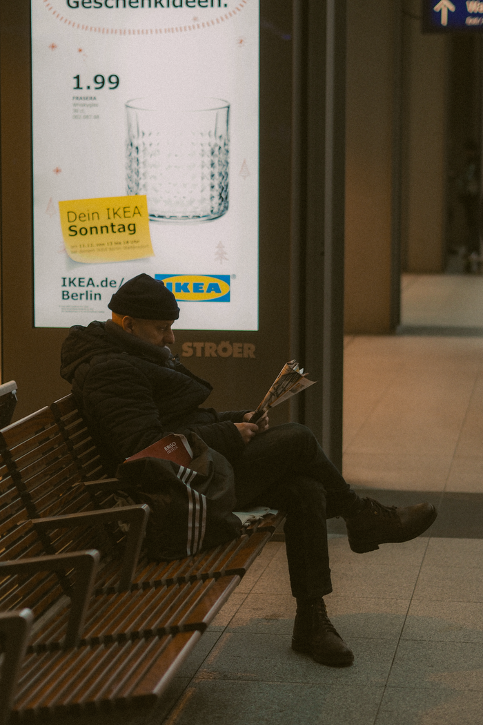 Adult man sitting and reading a newspaper beside an IKEA ad in urban Berlin setting.
